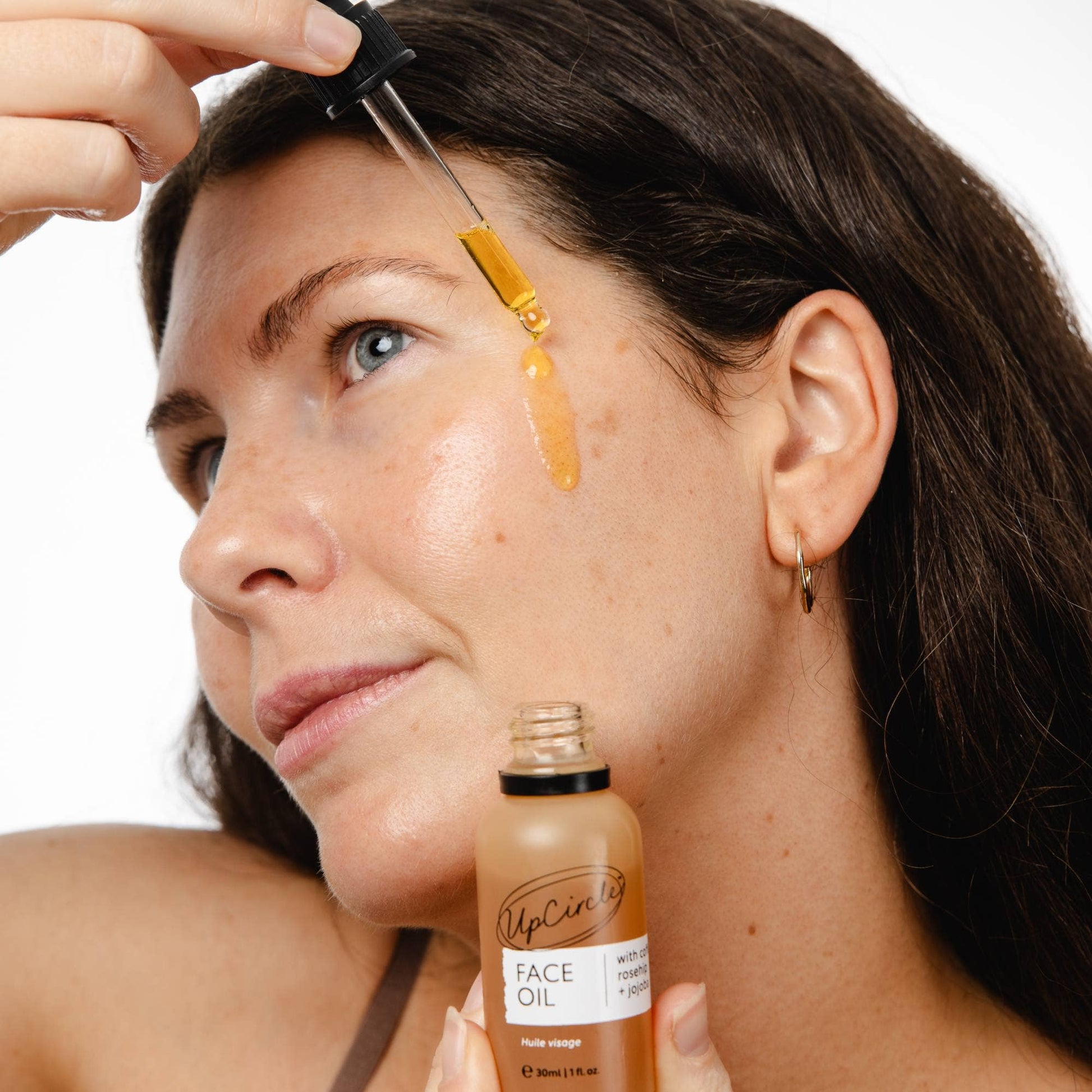 Woman applying face oil to her face with a dropper, holding a bottle labeled 'UpCycled Face Oil'.